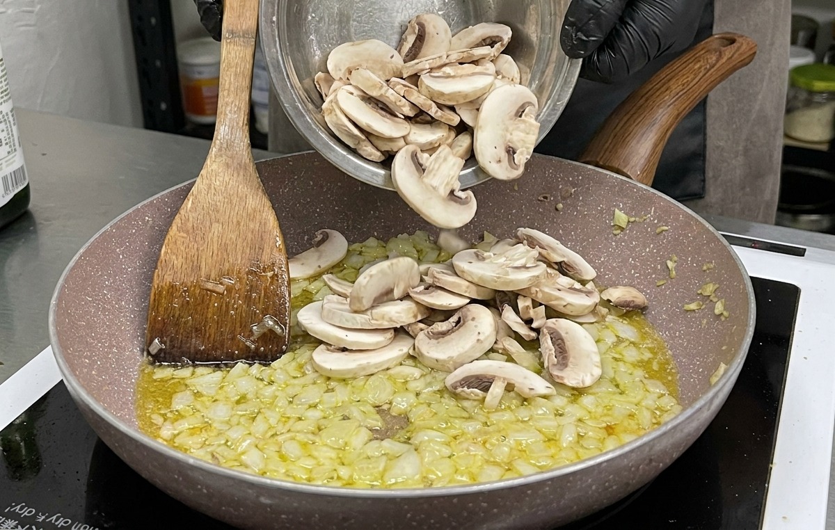 Una grande ciotola di funghi champignon bianchi crudi e affettati viene aggiunta alla padella con cipolle e aglio soffritti.