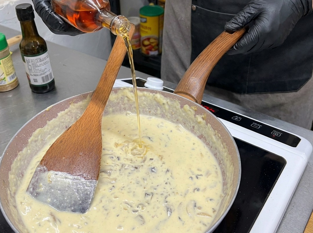 Una persona con guanti neri versa un liquido ambrato da una bottiglia di vetro in una padella colma di salsa ai funghi cremosa.