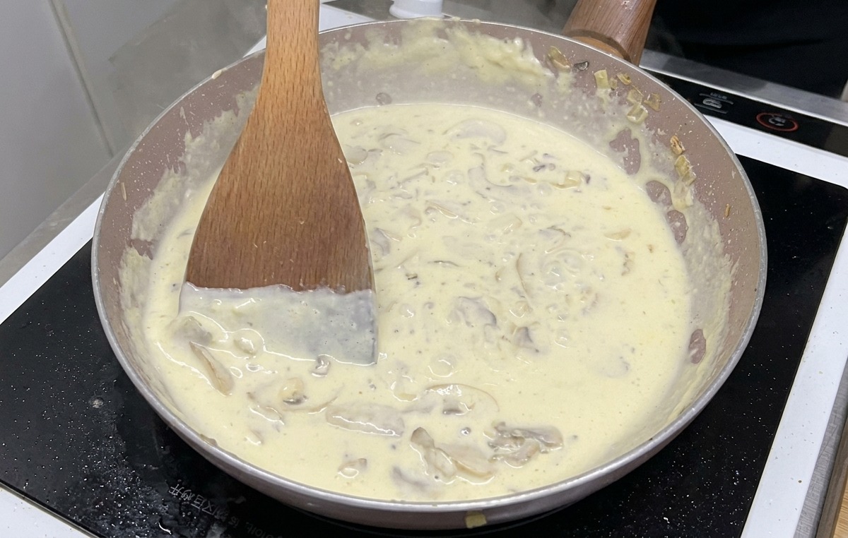 Una salsa ai funghi cremosa con fette di funghi visibili che bolle e sobbolle in padella mentre viene mescolata con una spatola di legno.