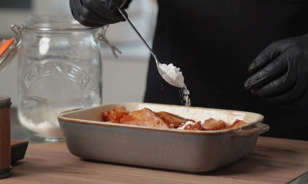 Una persona con guanti neri aggiunge un cucchiaio di amido di mais bianco a una pirofila rettangolare con le costine di maiale marinate.