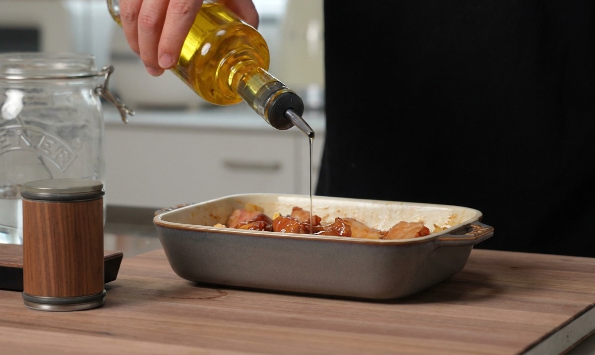 Olio dorato versato da una bottiglia di vetro su costine di maiale condite in una pirofila di ceramica grigia.