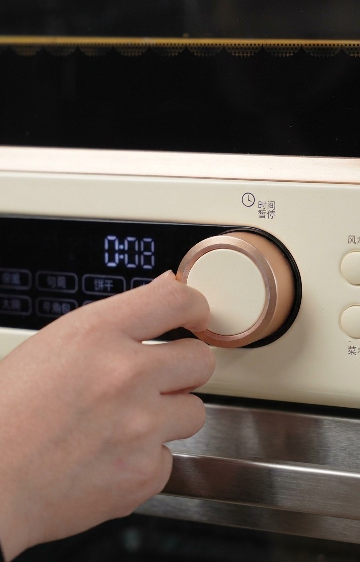 Una mano gira la manopola oro rosa di un forno color crema per impostare il tempo e la temperatura di cottura iniziali.