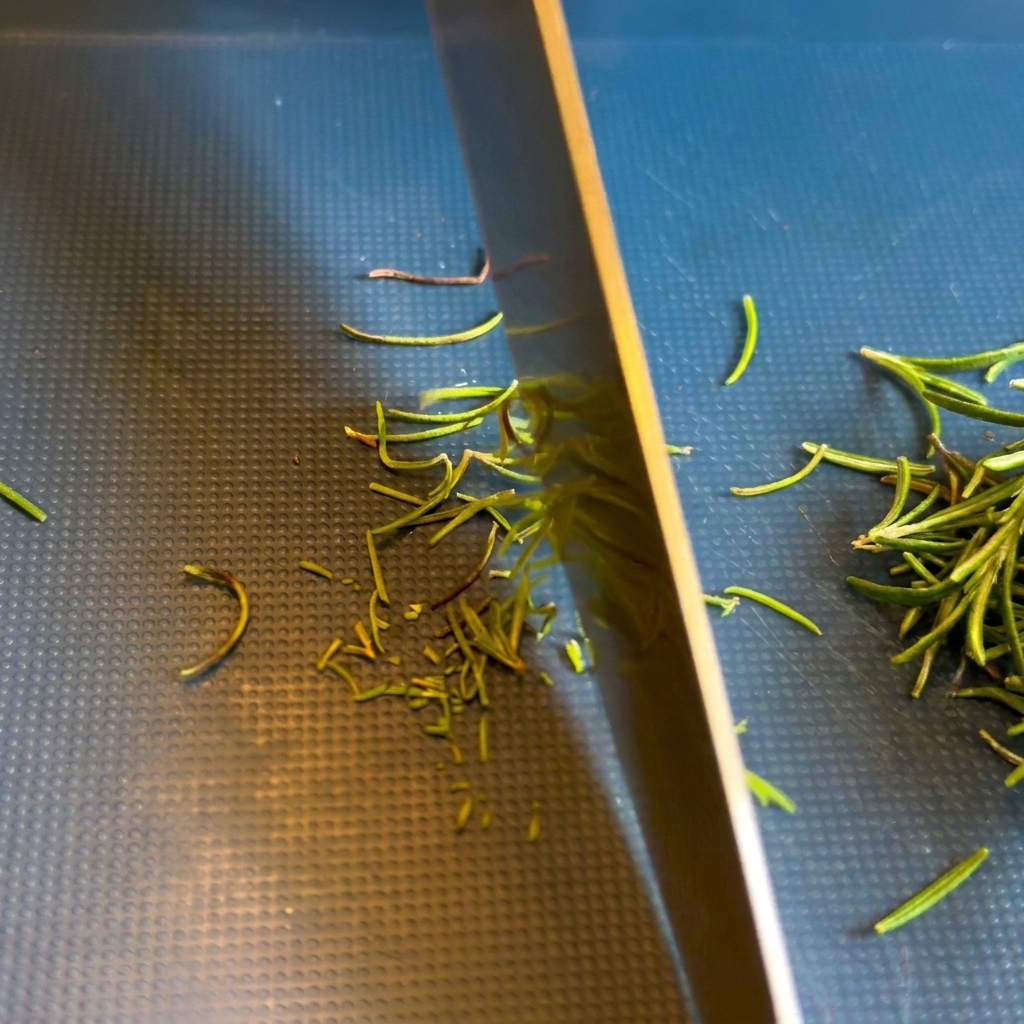 Primo piano della lama di un coltello da chef che trita aghi di rosmarino fresco su un tagliere scuro.