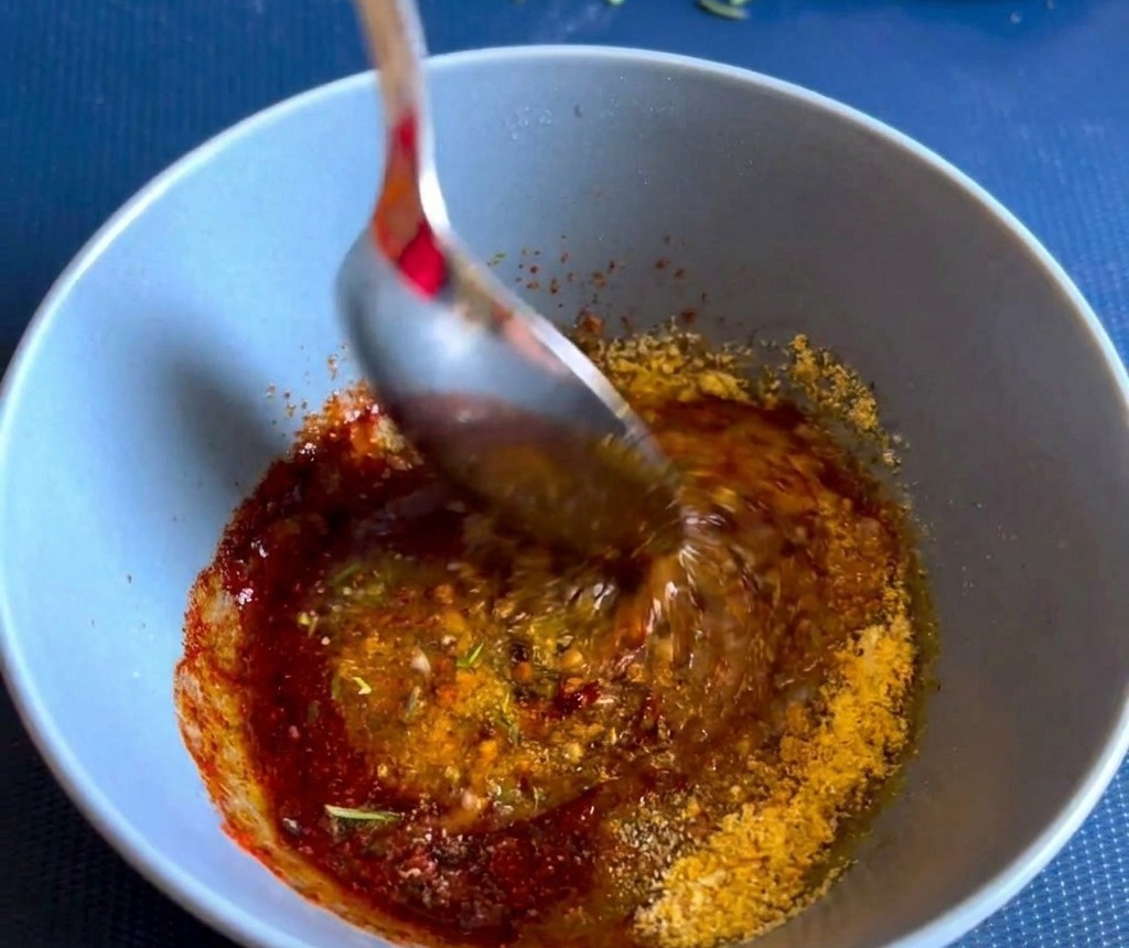 Un cucchiaio di metallo mescola una densa pasta di spezie ed erbe aromatiche rossastra allinterno di una ciotola di ceramica blu.
