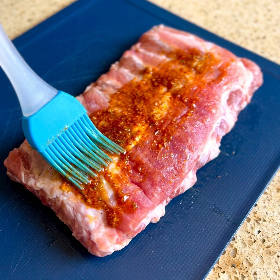 Un pennello in silicone blu distribuisce una densa marinata speziata sulla superficie di un pezzo di costine di maiale crude su un tagliere blu.