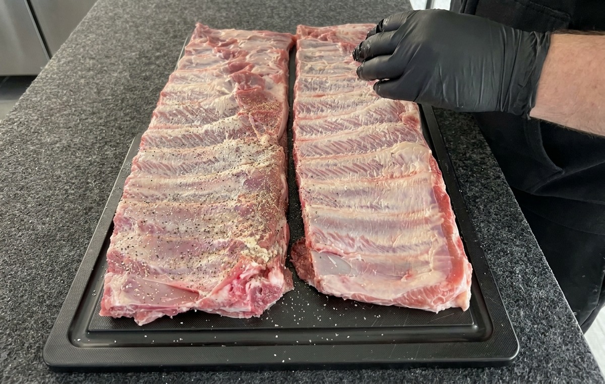 Due rack di costine di maiale crude condite con un rub secco di sale, pepe e aglio in polvere su un tagliere nero.