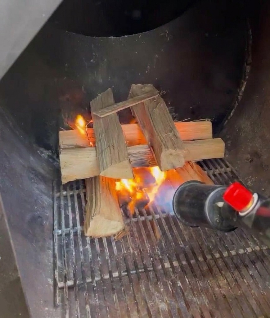 Un cannello a propano utilizzato per accendere una pila di ciocchi di legno di quercia allinterno del focolare metallico di un affumicatore.