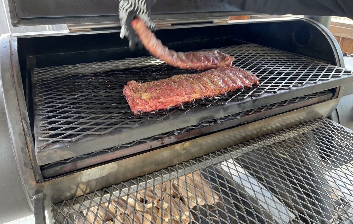 Una mano che indossa un guanto resistente al calore gira un rack di costine parzialmente affumicate sulla griglia metallica di un affumicatore.