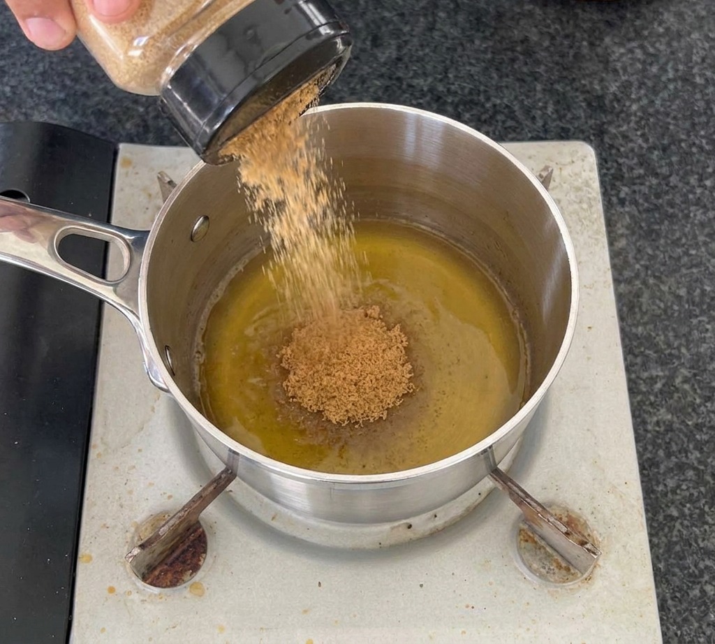Una mano versa dello zucchero di canna da un barattolo trasparente in un pentolino dacciaio contenente aceto di sidro di mele su un fornello.