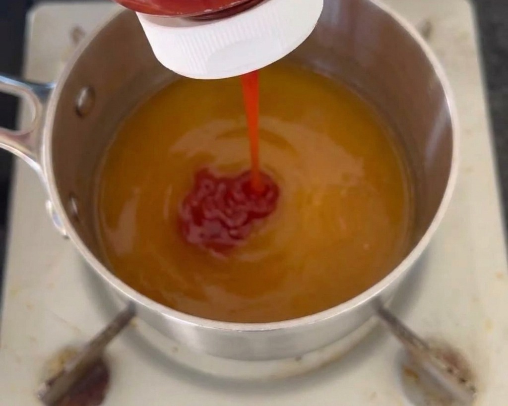 Un filo di ketchup rosso viene spremuto da una bottiglia in un pentolino contenente una base di salsa liquida giallastra.