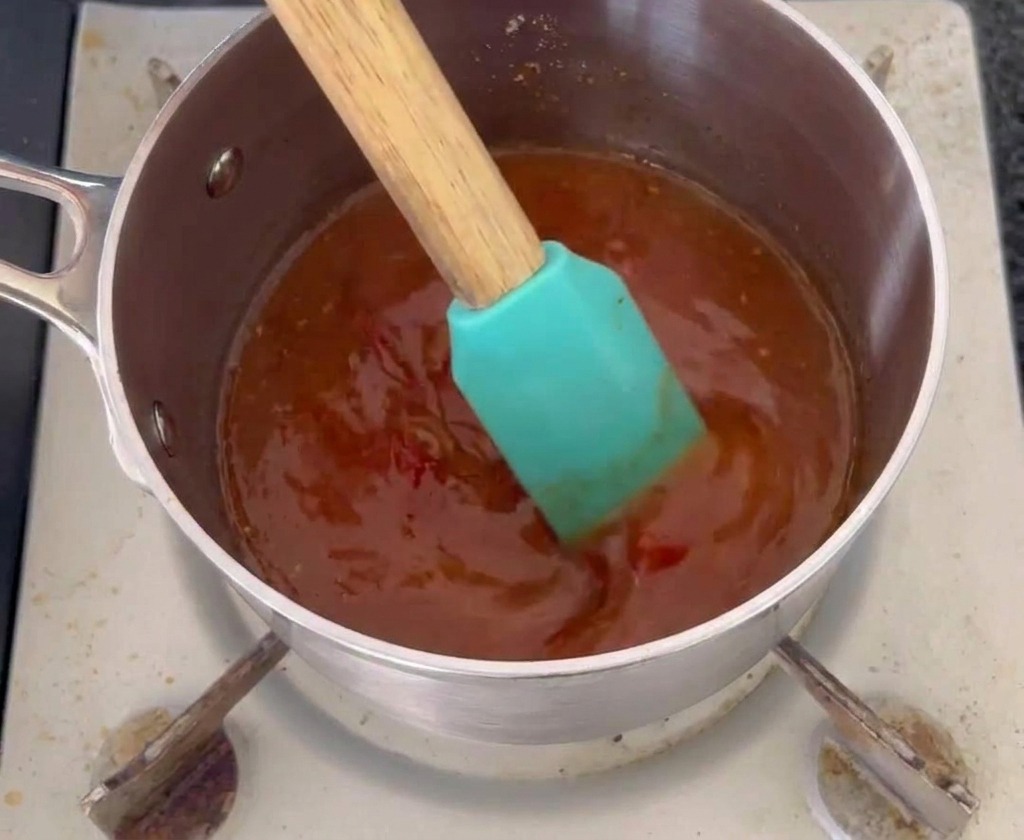 Una spatola in silicone color teal mescola una salsa mop arancione-rossastra scura in un piccolo pentolino di acciaio inossidabile.