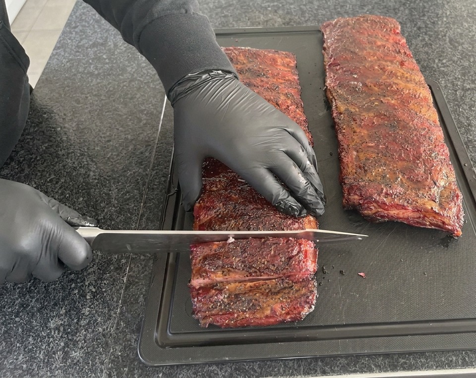 Una persona che indossa guanti in nitrile neri usa un grande coltello da chef per tagliare un lungo rack di costine affumicate rosso scuro su un tagliere nero.