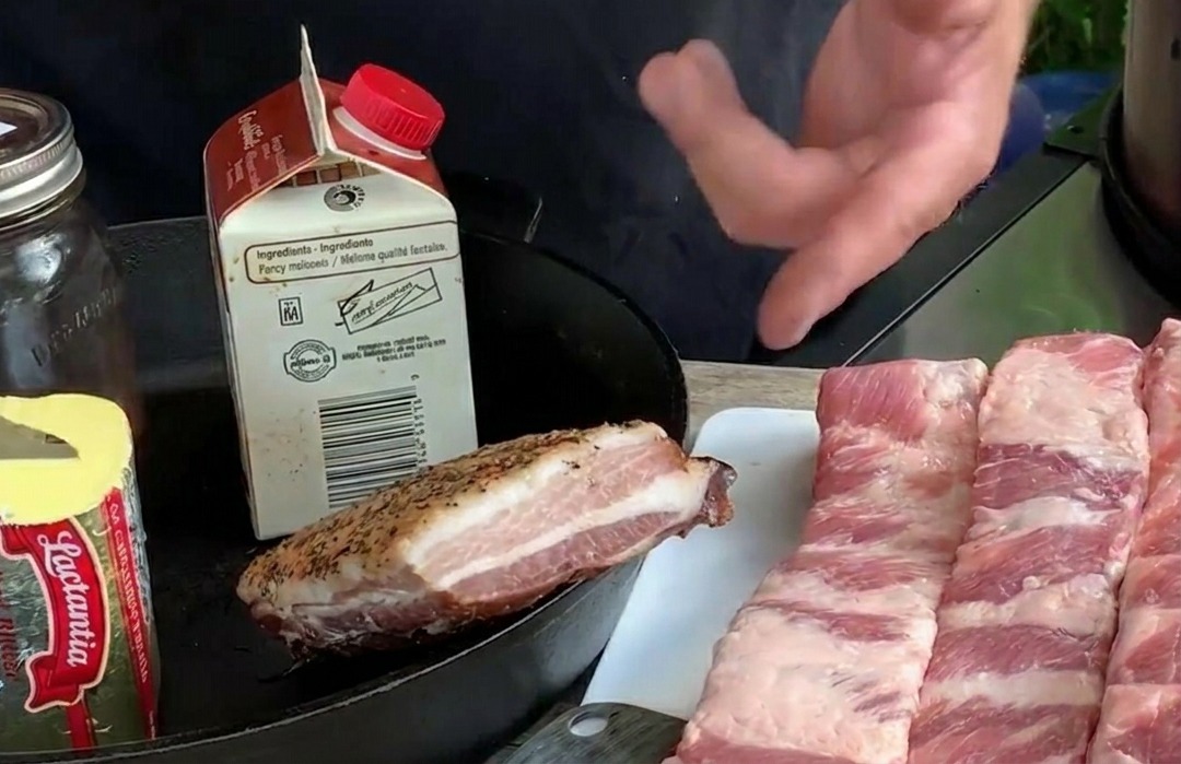 Diverse lunghe strisce di costine di maiale crude disposte su un tagliere bianco insieme a ingredienti come burro e mirtilli freschi.