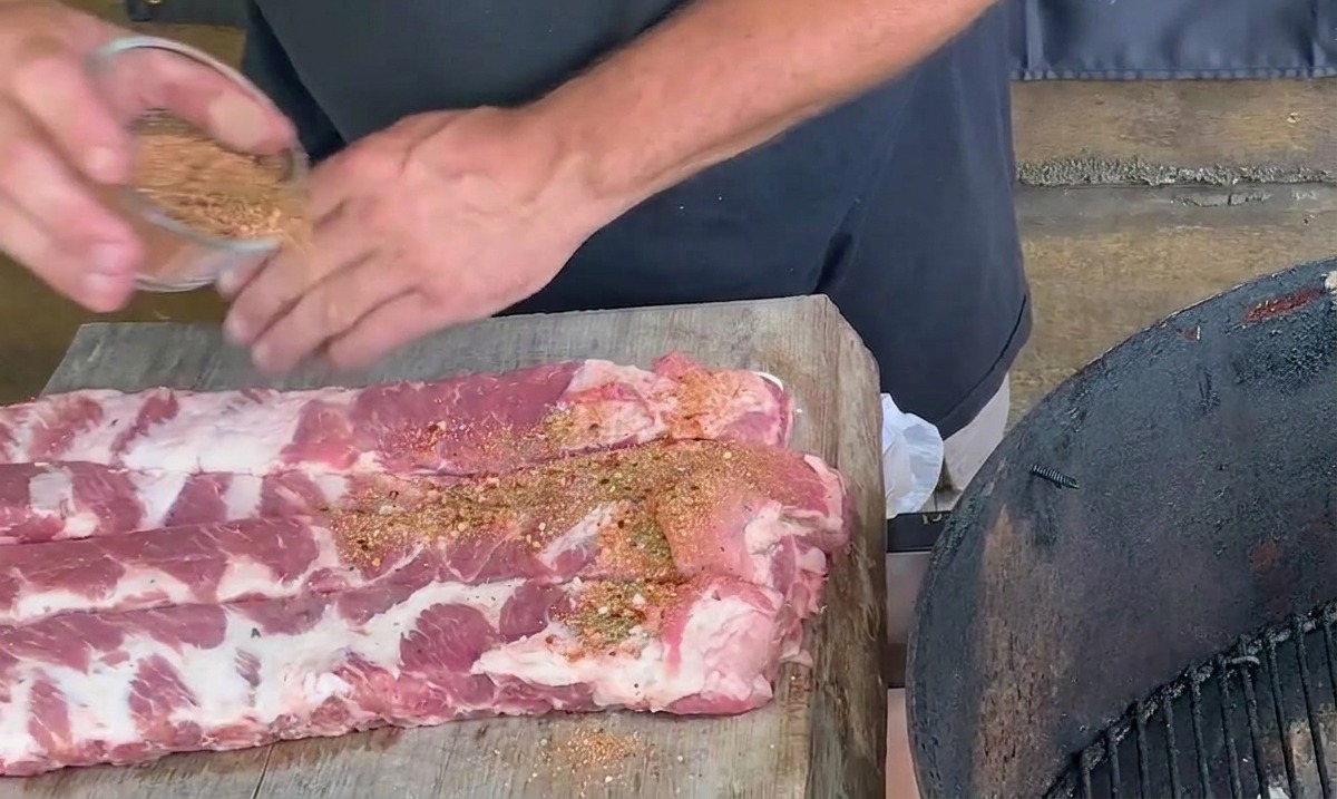 Mani che cospargono una miscela di spezie marrone-rossastra su costine di maiale crude disposte su un tagliere di legno.