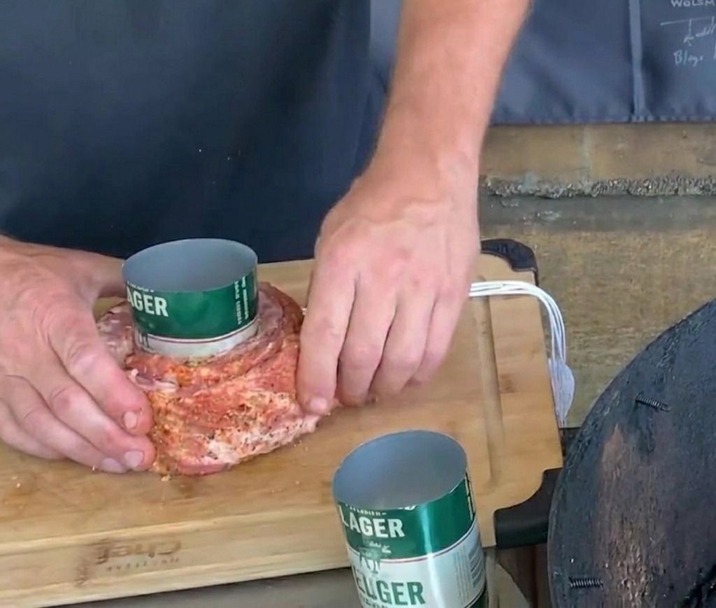 Mani che legano con cura dello spago bianco attorno a una costina di maiale condita e avvolta attorno a una lattina di birra verde.