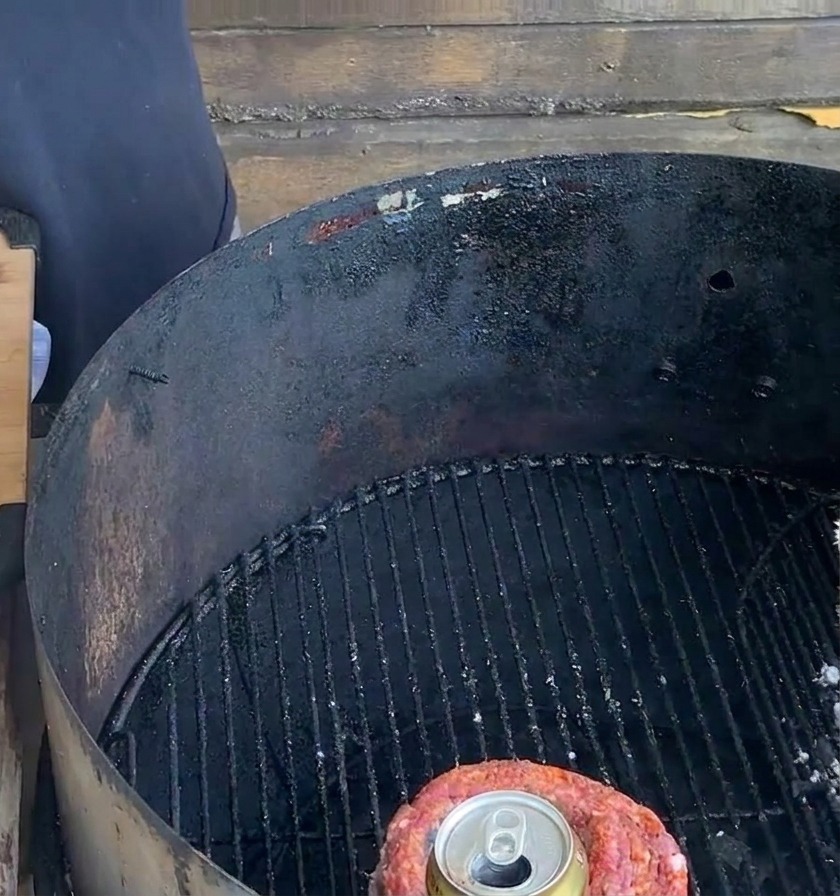 Un cestino di carne legato contenente una lattina di birra si trova sulla griglia di un barbecue a carbone nero, accanto a un tagliere in legno.