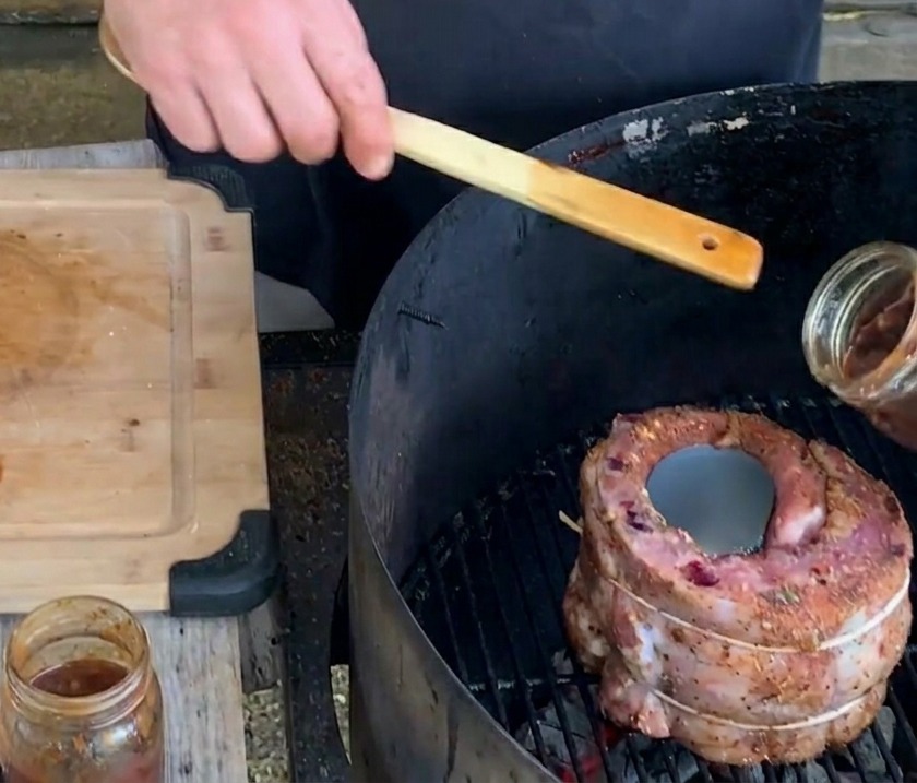 Una mano versa una densa e scura salsa ai mirtilli da un barattolo di vetro in una lattina di birra posizionata al centro di un cestino di costine sulla griglia.
