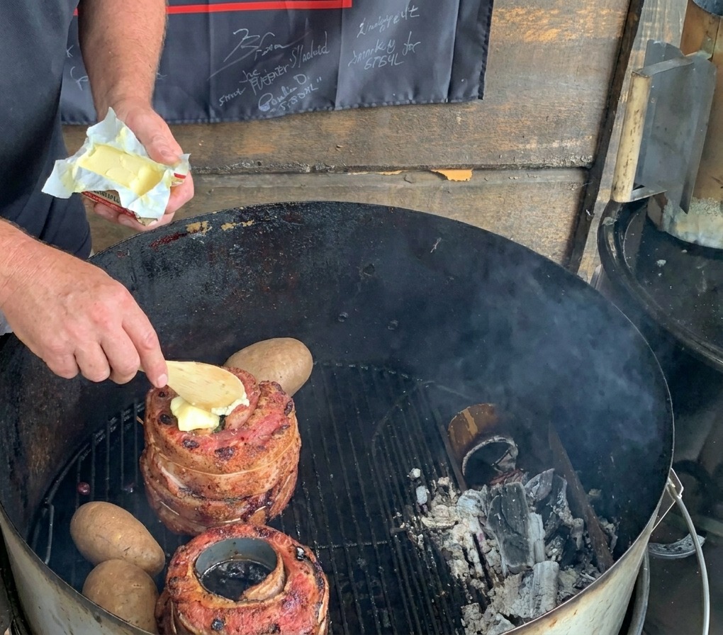 Un cucchiaio di legno posiziona una noce di burro sulla carne su un barbecue a carbone accanto a patate intere.