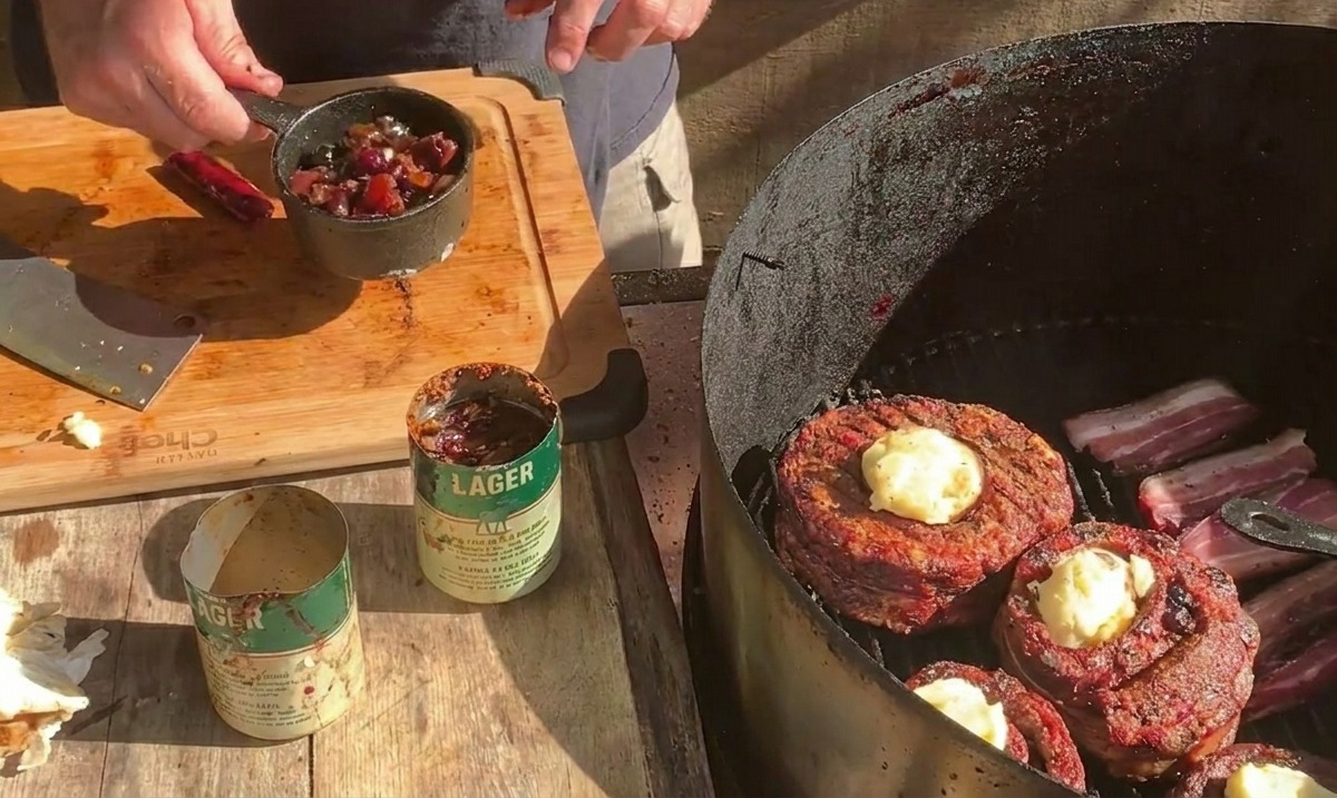 Una persona tiene un piccolo pentolino di metallo pieno di salsa ai mirtilli sopra un barbecue dove i cestini di carne sono ricoperti di formaggio.