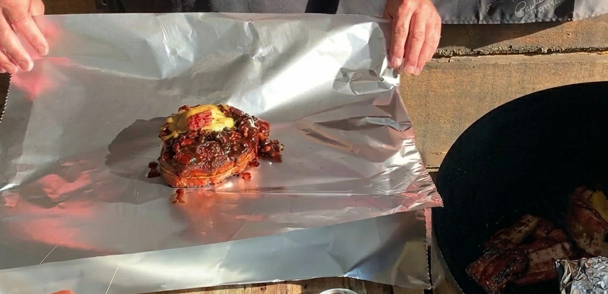 Un cestino di costine BBQ pronto, sormontato da formaggio fuso, posizionato su un grande foglio di alluminio pronto per essere avvolto.