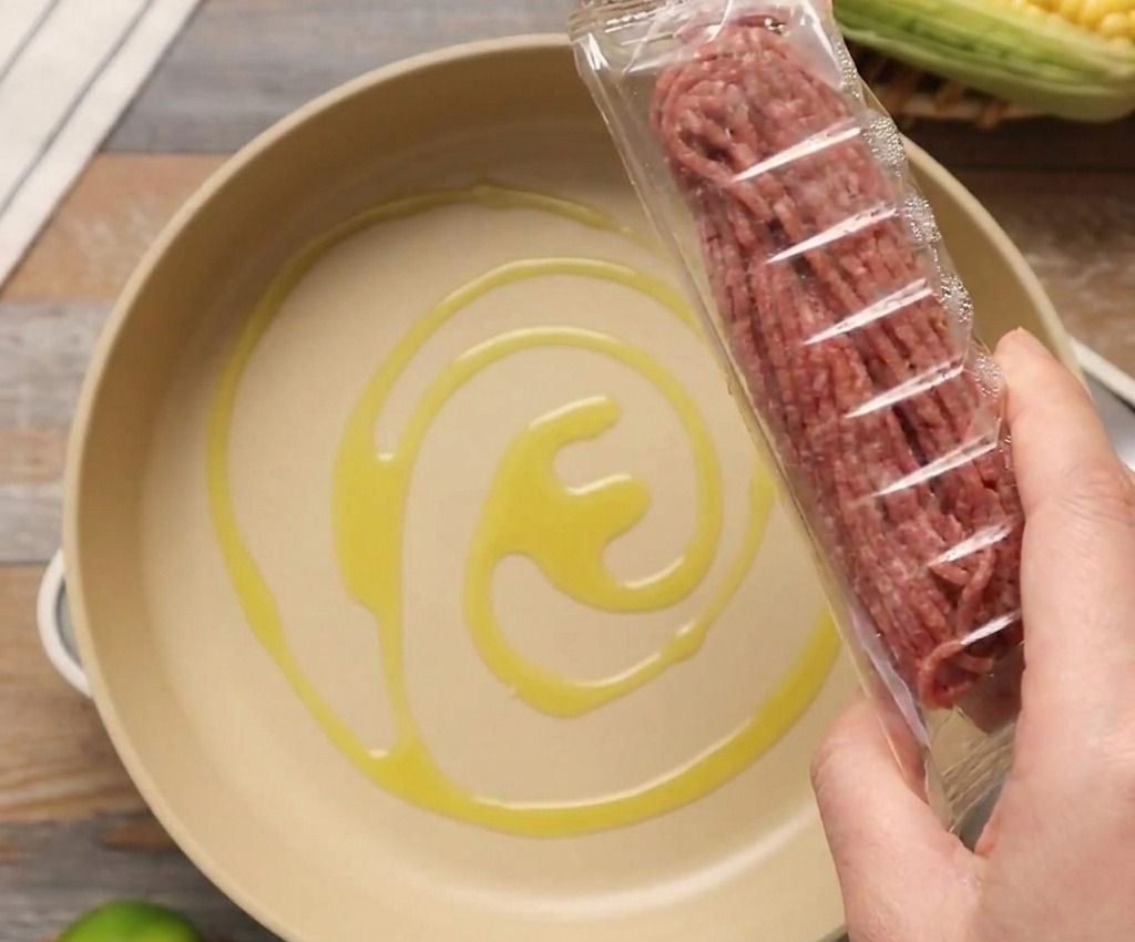 Un pacchetto di carne macinata cruda tenuto sopra una padella chiara contenente un filo dolio doliva.