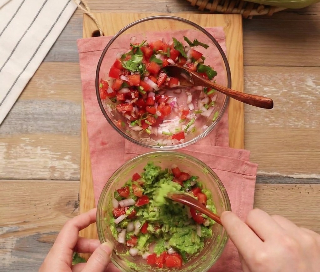 Una persona usa un cucchiaio di legno per incorporare della salsa di pomodoro fresca in una ciotola di avocado schiacciato.
