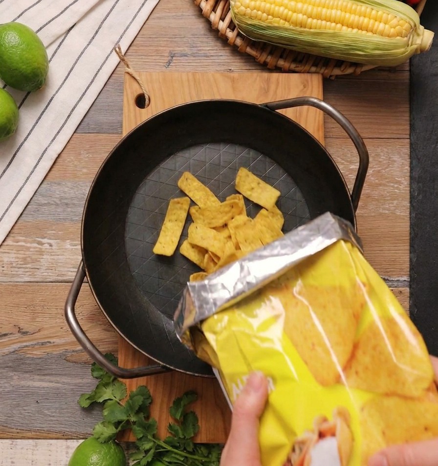 Delle tortilla chips triangolari gialle vengono versate da un sacchetto in una pesante padella di ghisa nera su una superficie di legno.