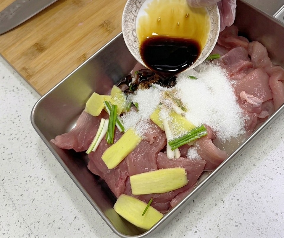 Condimento liquido scuro versato da una ciotolina su fette di maiale crudo, zenzero e cipollotti in una teglia.