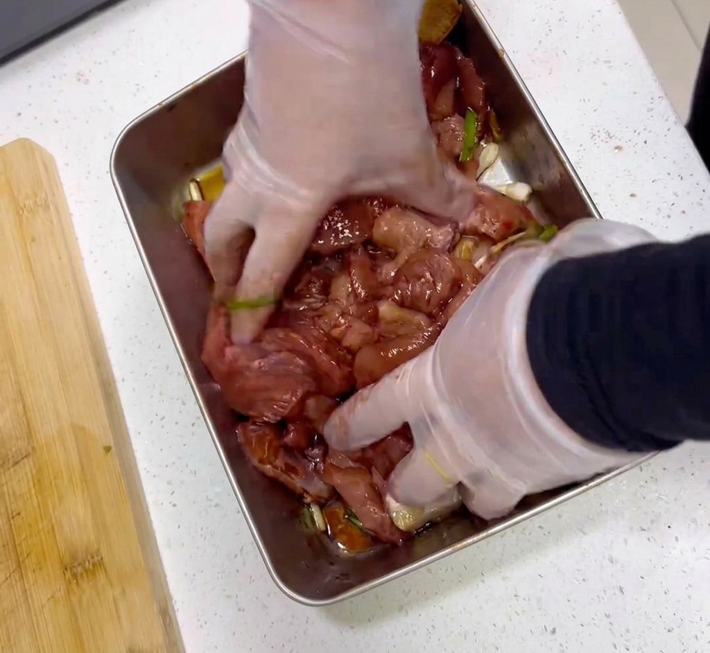 Mani con guanti di plastica trasparenti che mescolano fette di carne cruda con una marinata rosso scuro in un vassoio dacciaio.