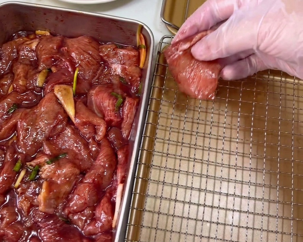 Una mano in un guanto di plastica trasparente che adagia una fetta di maiale marinato rosso scuro su una griglia color oro.