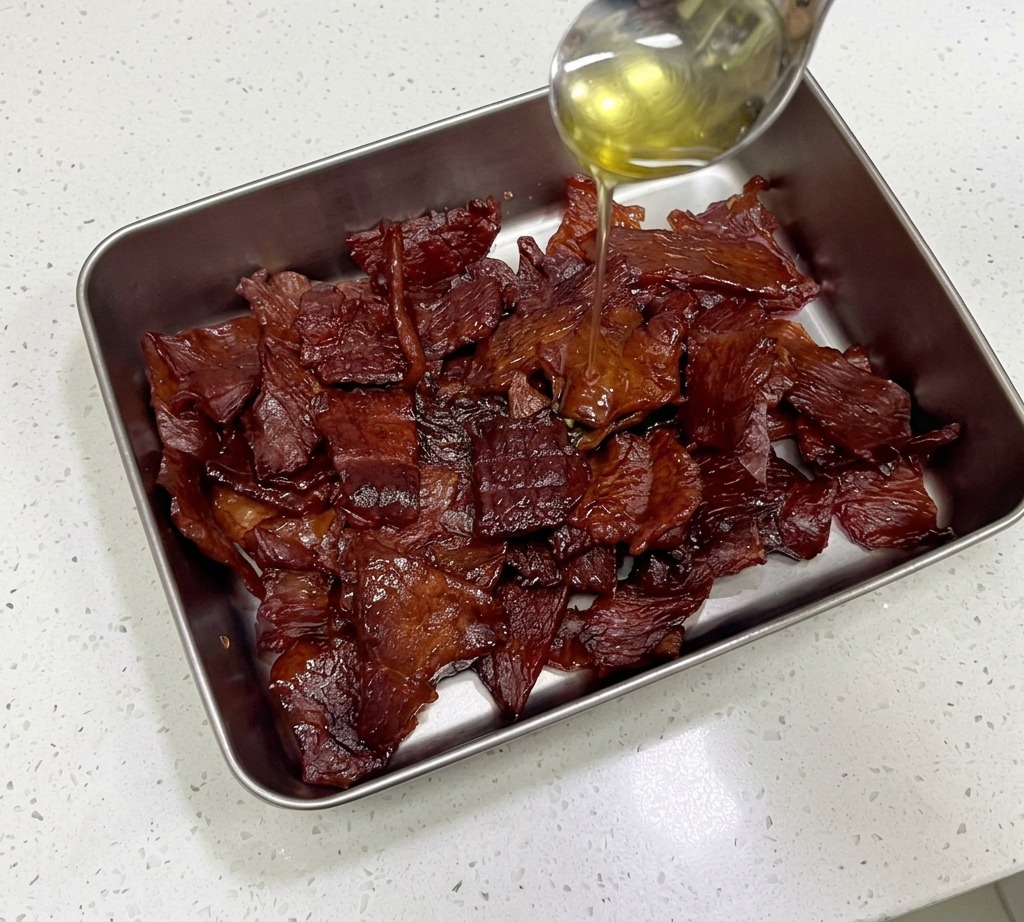 Miele dorato versato da un cucchiaio dacciaio su una pila di strisce di carne secca rosso mogano lucido in un contenitore metallico.