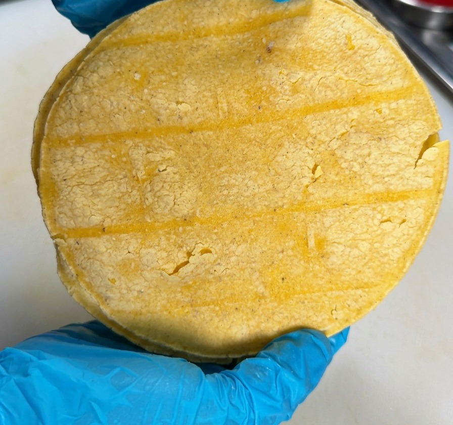 Una persona con guanti blu tiene in mano una pila rotonda di tortillas di mais giallo sopra un piano di lavoro bianco.