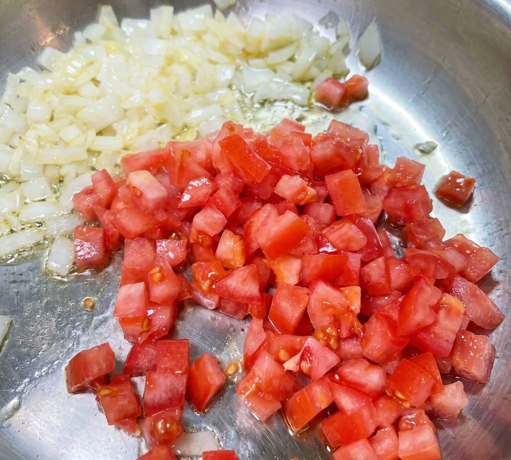 Pomodori rossi a cubetti mescolati in una padella insieme a cipolle soffritte traslucide.