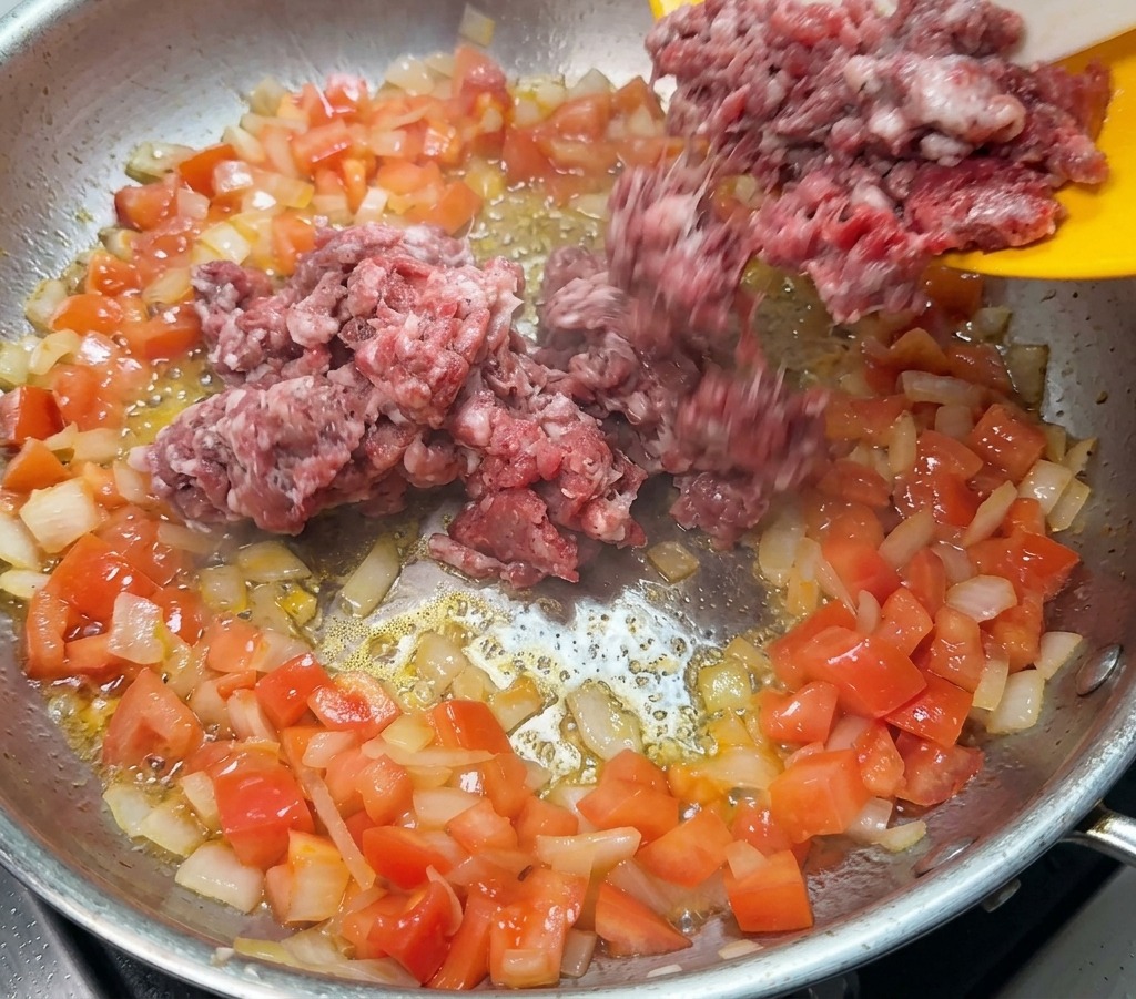 Carne macinata cruda che viene aggiunta in padella sopra il soffritto di cipolle e pomodori.