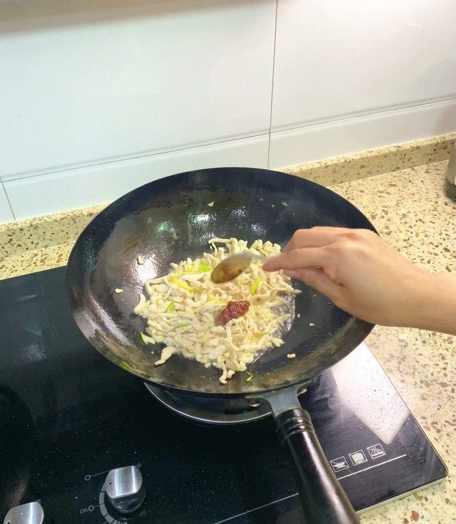 Un cucchiaio dacciaio che versa una generosa porzione di densa pasta di fave Pixian rossa nel wok con il pollo e gli aromi.