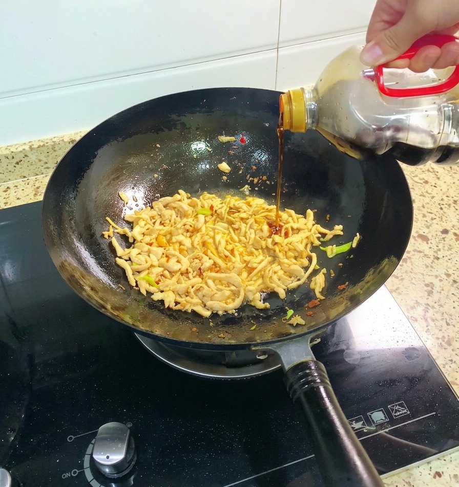 Una mano che versa la salsa di soia scura direttamente dalla bottiglia nel wok con il pollo e il condimento speziato.
