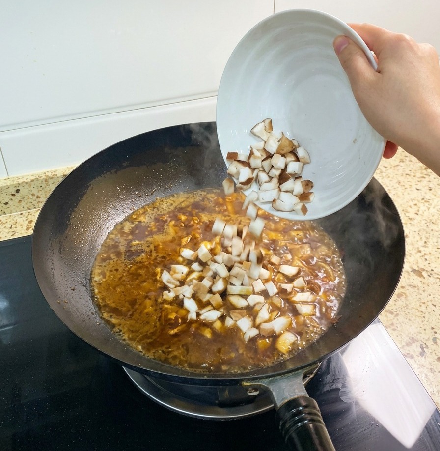 Una manciata di funghi shiitake tagliati a dadini che vengono tuffati nel brodo rossastro che sobbolle nel wok.