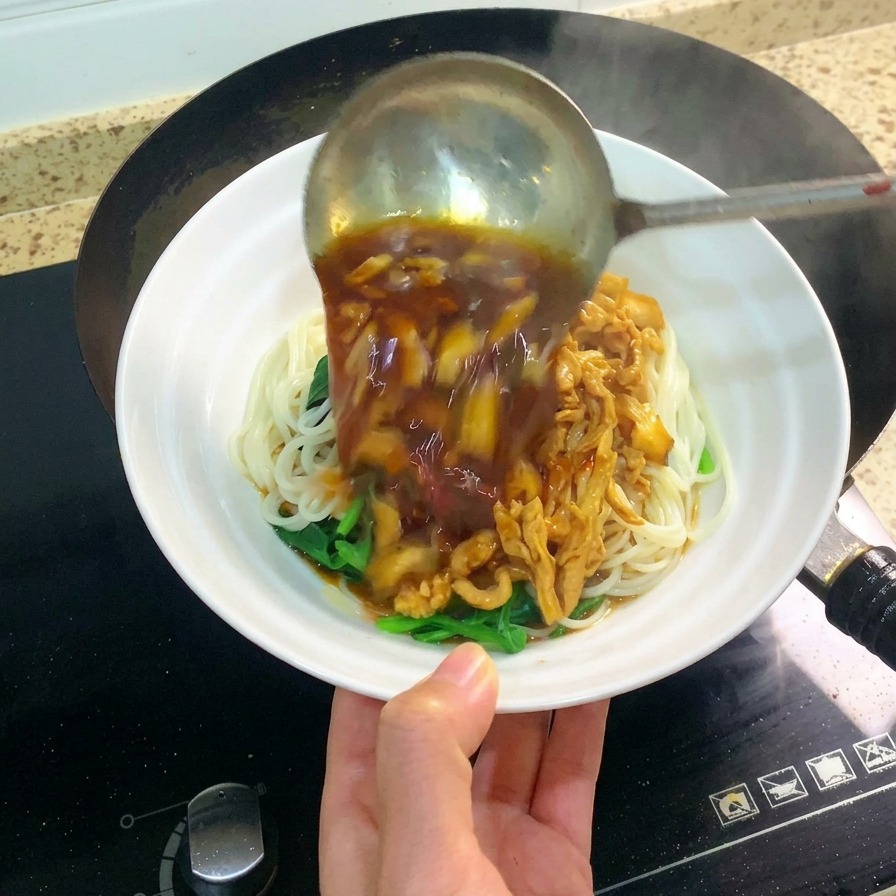 Un grande mestolo di metallo che versa il condimento scuro e saporito di pollo e funghi su una ciotola di noodles bianchi.