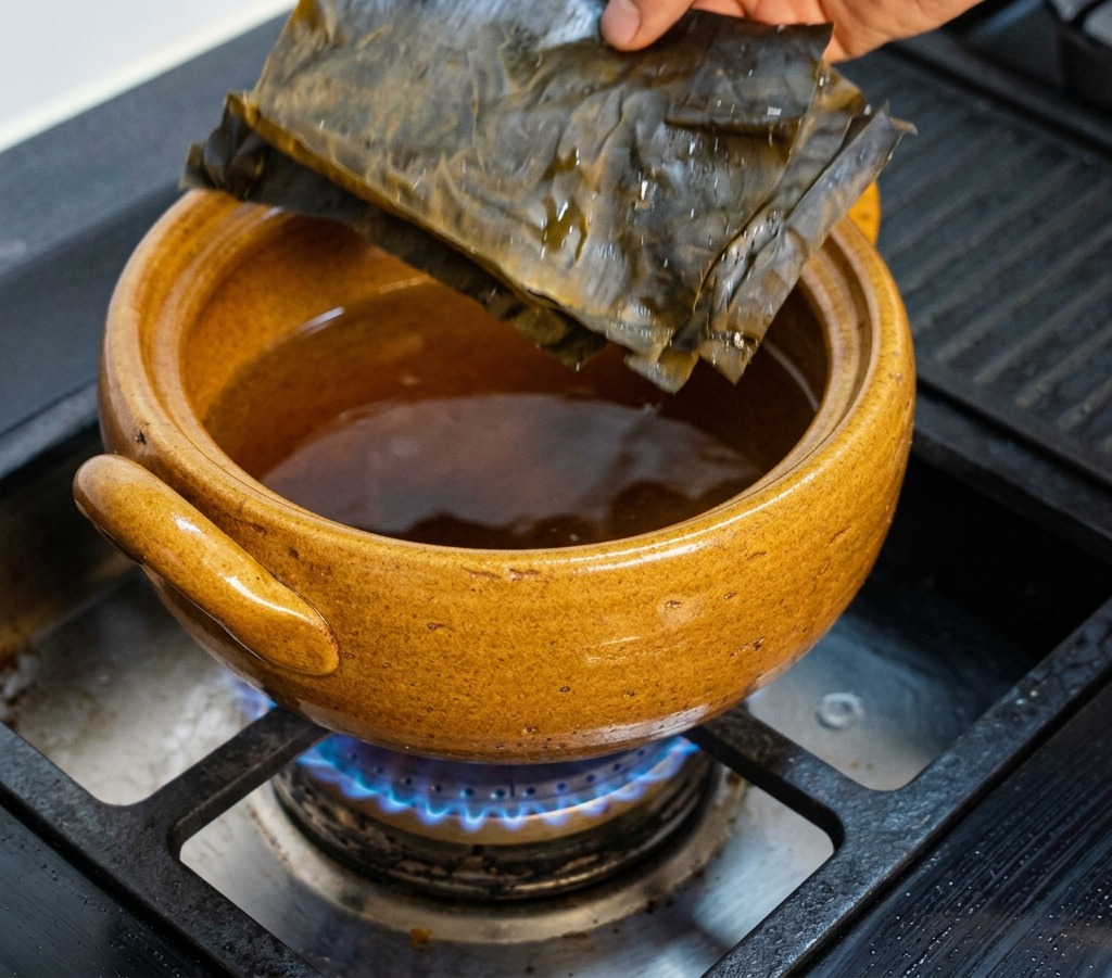 Una mano che adagia fogli rettangolari di alga kombu verde scuro in una pentola di terracotta marrone su un fornello a gas.