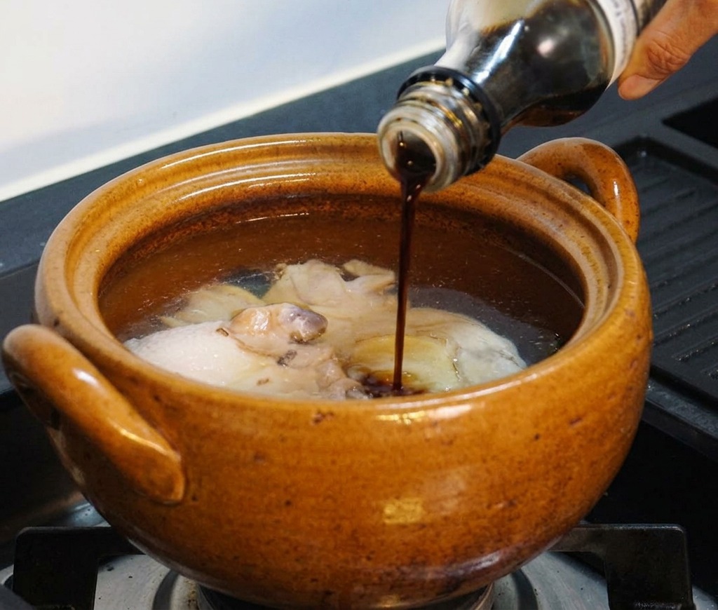 Una mano che versa del condimento liquido scuro da una bottiglia di vetro nella pentola con il pollo e lacqua.