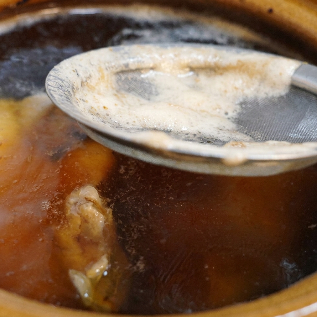 Un colino metallico a maglia fine che raccoglie la schiuma bianca dalla superficie di un brodo di pollo scuro e sobbollente in una pentola di ceramica.
