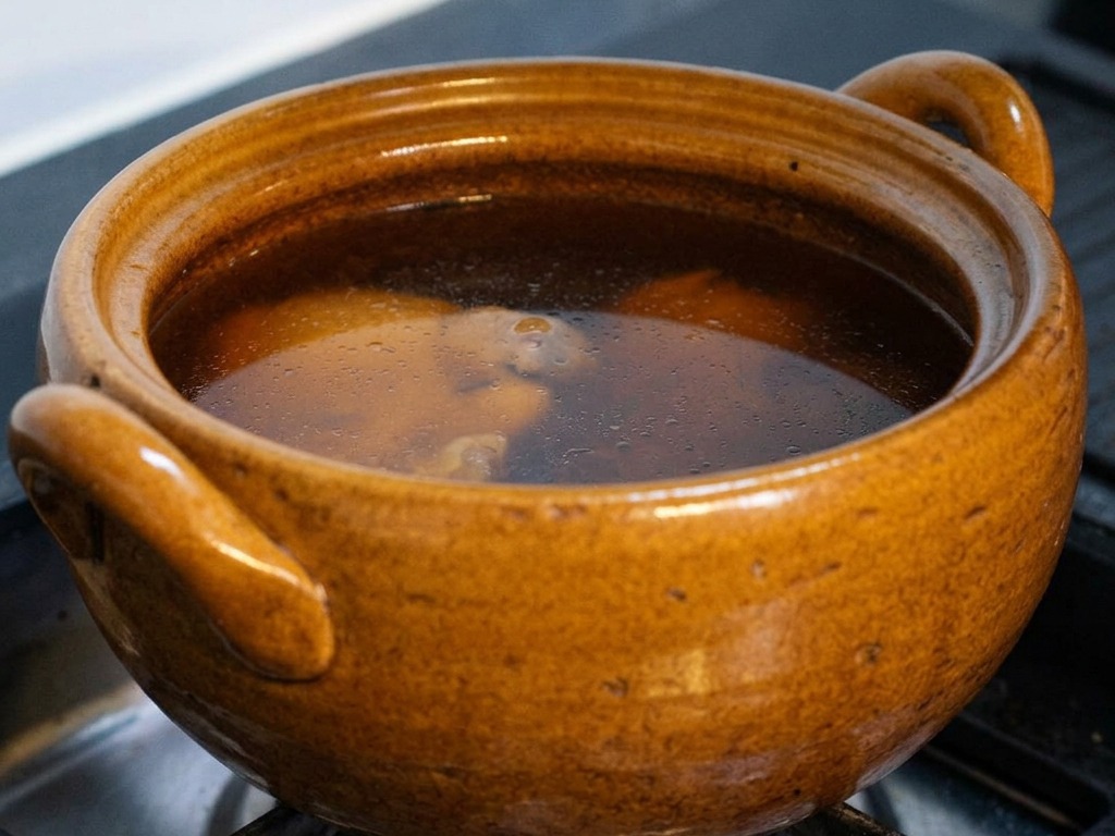 Una pentola di ceramica marrone su un fornello a gas piena di brodo scuro alla soia che sobbolle dolcemente, con il coperchio parzialmente sollevato per mostrare il pollo e la kombu allinterno.