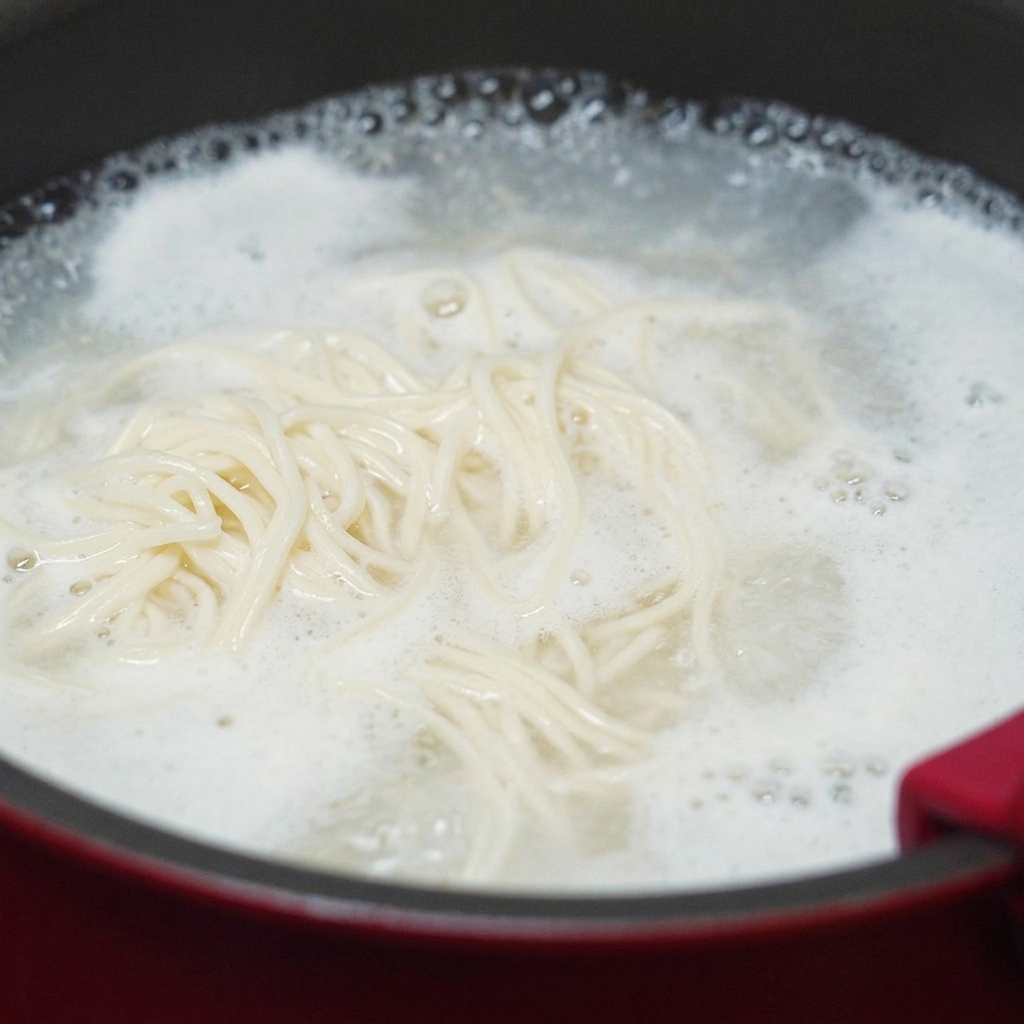 Noodles bianchi di frumento che girano in una pentola dacqua in pieno bollore, con una densa schiuma di amido che sale verso i bordi.