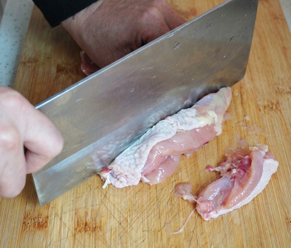 Una persona usa una grossa mannaia di metallo per disossare una coscia di pollo cruda su un tagliere di legno.