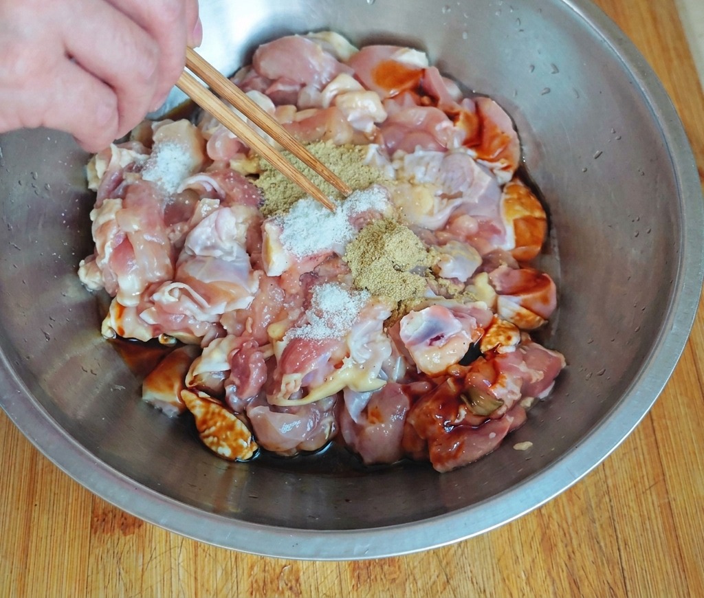 Una mano usa le bacchette per cospargere del condimento chiaro su pezzi di pollo crudo in una ciotola di acciaio inox con salse scure.