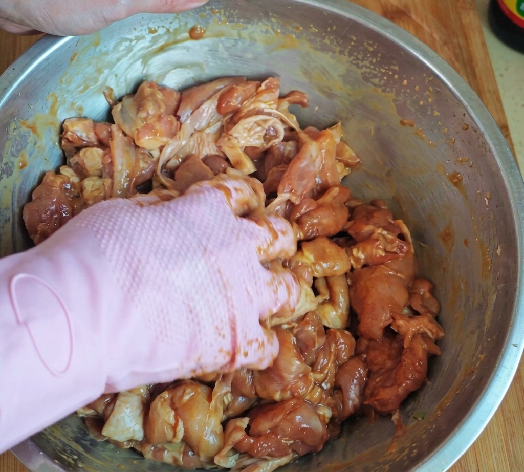 Una mano con un guanto rosa massaggia accuratamente i pezzi di pollo marinati, mescolandoli con amido e olio in una grossa ciotola dacciaio.