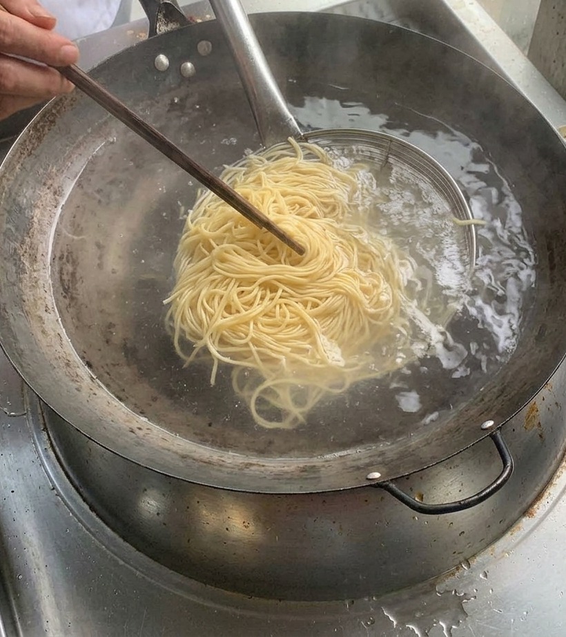 Noodles cotti allinterno di un colino metallico sollevato sopra una pentola di acqua bollente.
