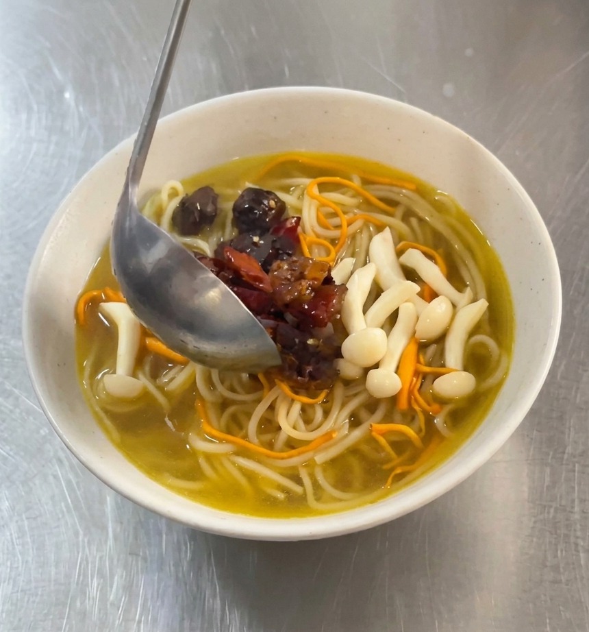 Un mestolo di metallo versa pezzetti di pollo marinato piccante rosso e peperoncini secchi su una ciotola di noodles bianchi in brodo dorato.