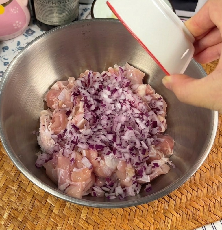 Una mano versa una ciotolina di cipolle rosse tritate e aglio sminuzzato sui pezzi di pollo crudo in una ciotola di metallo.