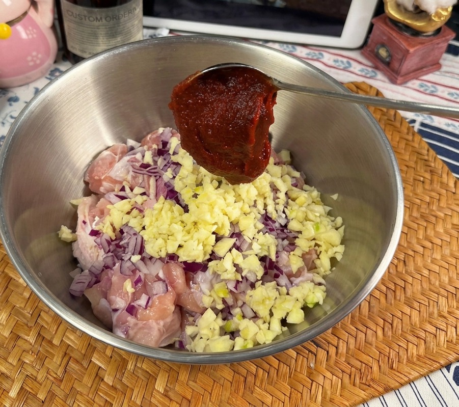 Un cucchiaio di metallo raccoglie una generosa porzione di densa pasta rossa Gochujang sopra una ciotola con pollo crudo, aglio e cipolle.